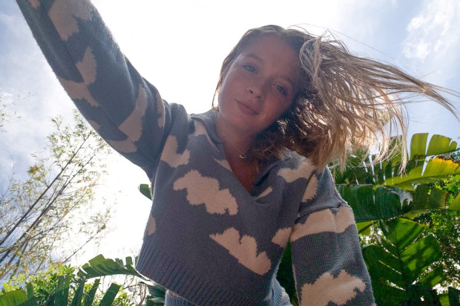 A person with long blonde hair is shot from a low angle. They are wearing a blue knitted jumper with clouds on it, and behind them the sky is blue. There are also bright green plants behind the person.