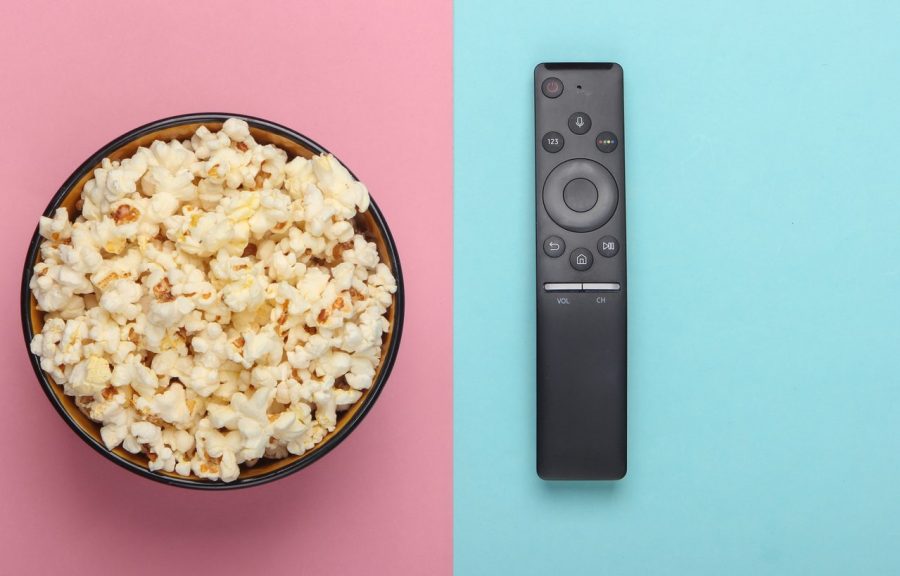 A bowl of popcorn and a slim, black TV remote are viewed from above. The background is split in two slightly left of centre. The left hand side is pink pastel, and the right hand side is blue pastel.