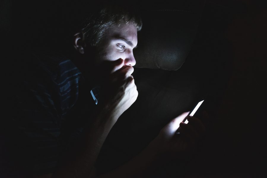 A man's face is partially illuminated as he scrolls on his phone in the dark