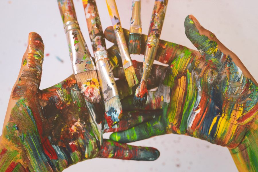 A pair of hands covered in multicoloured paint hold paintbrushes also stained with colour