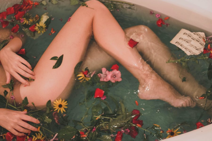 Two legs are seen in a bathtub. There are flowers in the bath water, and you can only see the person's legs and hands.