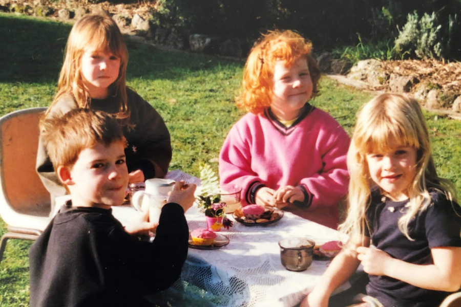 An image of four small children looking at the camera. They appear to be at a picnic table, with small cupcakes and cups and saucers on the white lace tablecloth. They are outside - behind them is green grass