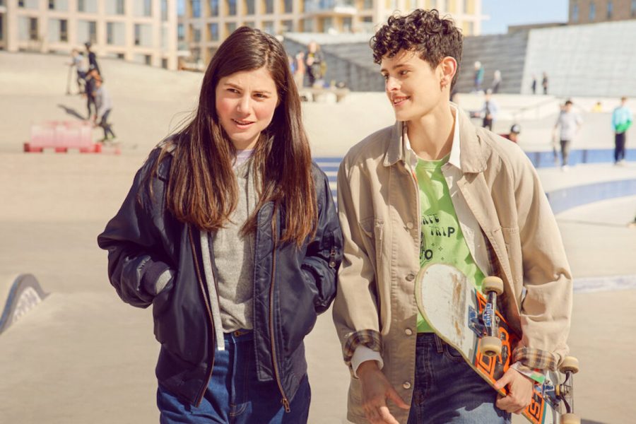 A still from Anne+, a Netflix film. There are two people walking in a skatepark, half-smiling. The person on the left has long dark hair and a denim jacket. On the right, the person has short curly hair, a green t-shirt and is holding a skateboard, looking at the person on the left.