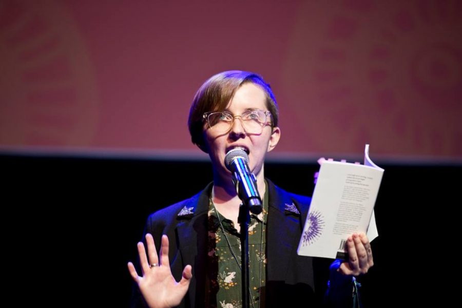 An image of a person with short red hair and glasses at a microphone, talking into it. they're holding a book and looking upwards.