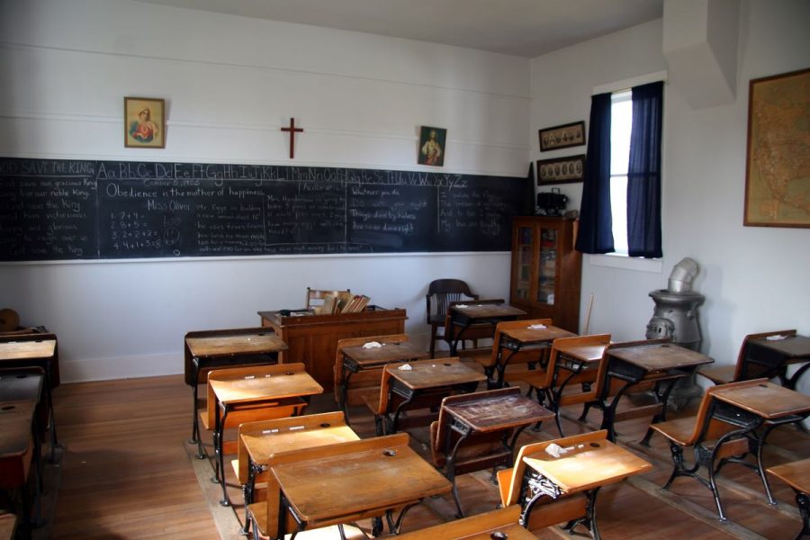 The setting of a vintage-looking Christian school. Old wooden student desks line the room, with the front of the room a blackboard with white chalk on it, a wooden cross and two religious images in frames hanging on the wall.