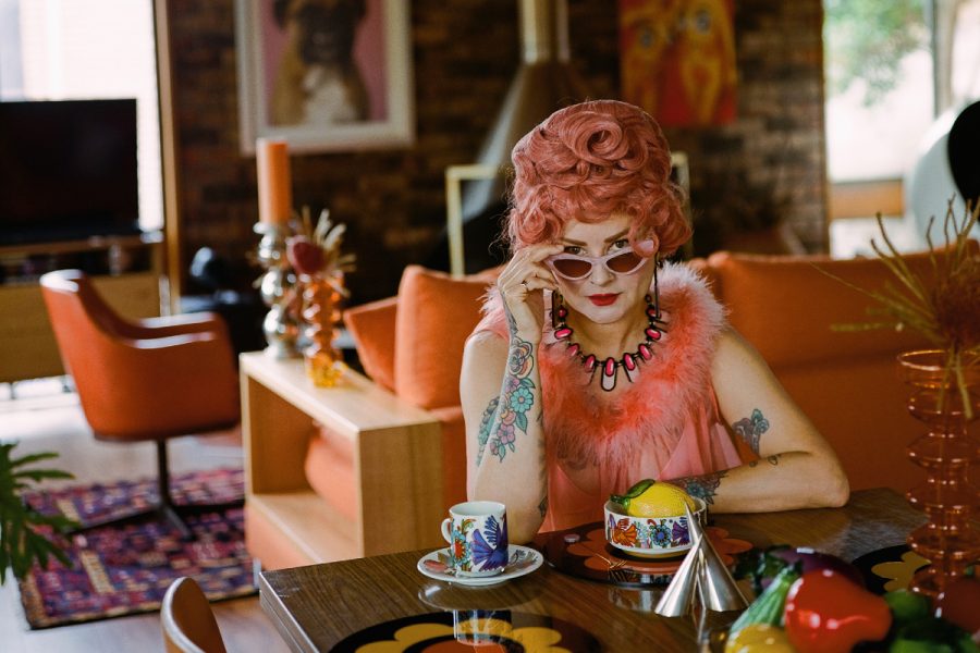 A person with a pink beehive hairstyle, tattoos and funky jewellery sitting at a table in a retro-styled room. Taken by Liz Ham.