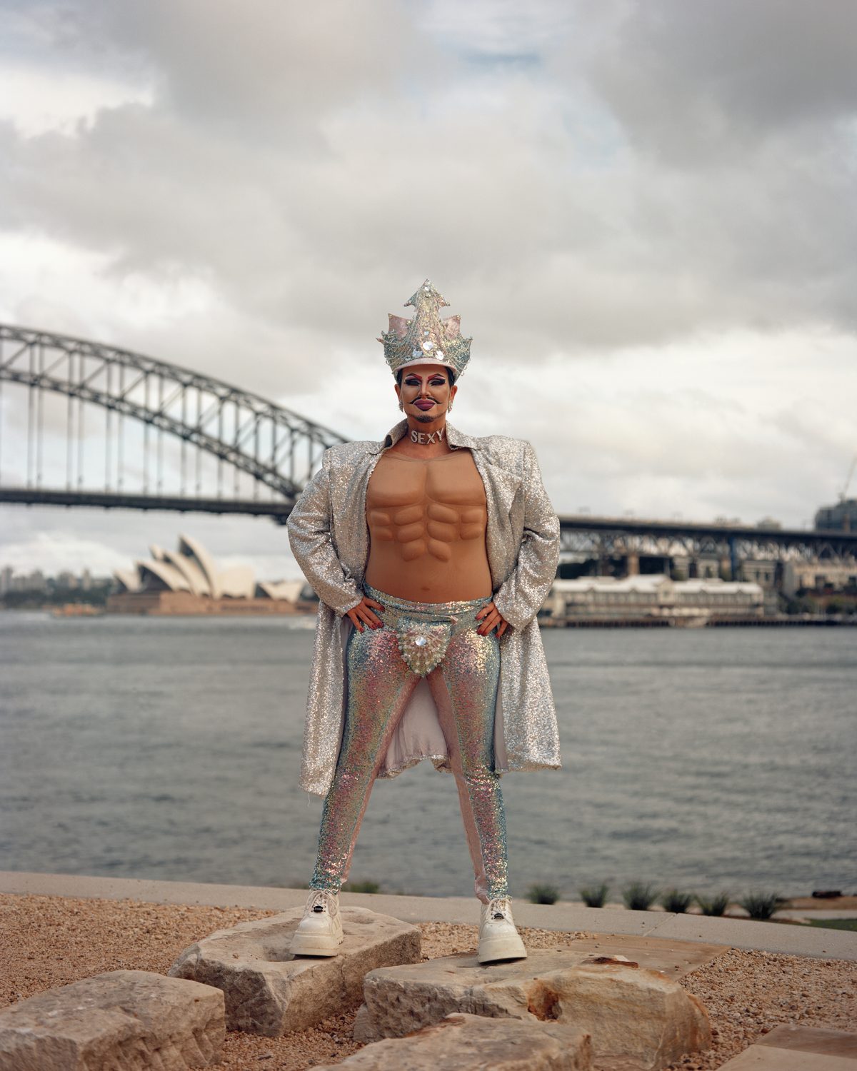 A person standing in a glittery, draggy outfit and chestpiece in front of the Sydney Harbour Bridge.