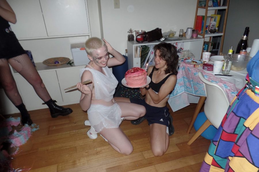 Two people crouch on the ground, smiling holding a birthday cake. They appear to be at a party in the kitchen. This is the image for the article Queer Platonic Intimacy