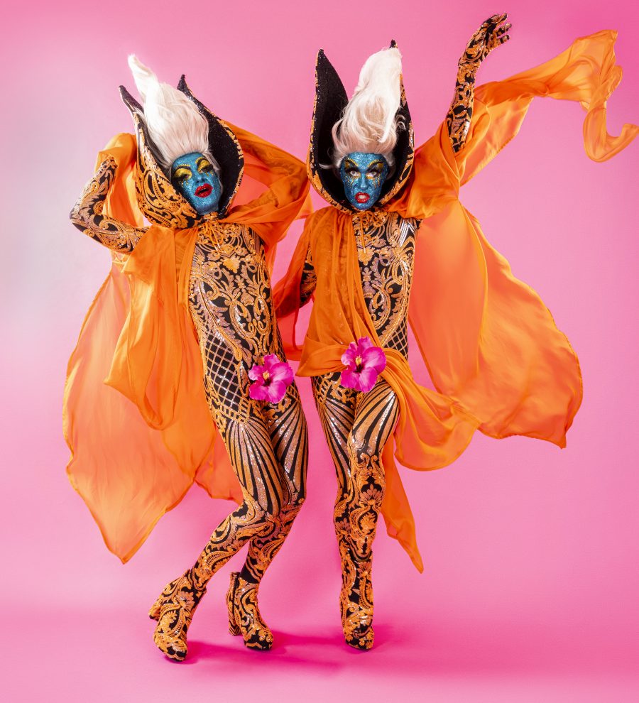 The Huxleys striking a pose with glittery blue faces and red lips, orange capes and beehive wigs. They have gold adorned jumpsuits and flowers on their crotches.