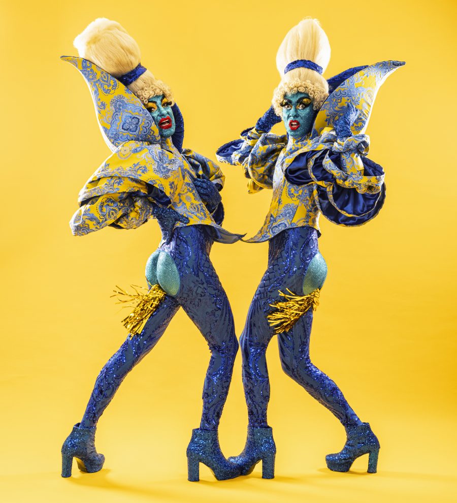 The Huxleys wear Leigh Bowery-inspired patterns in blue and yellow, beehive wigs and glittery bodysuits. Their bums are exposed and gold streamers are coming out.