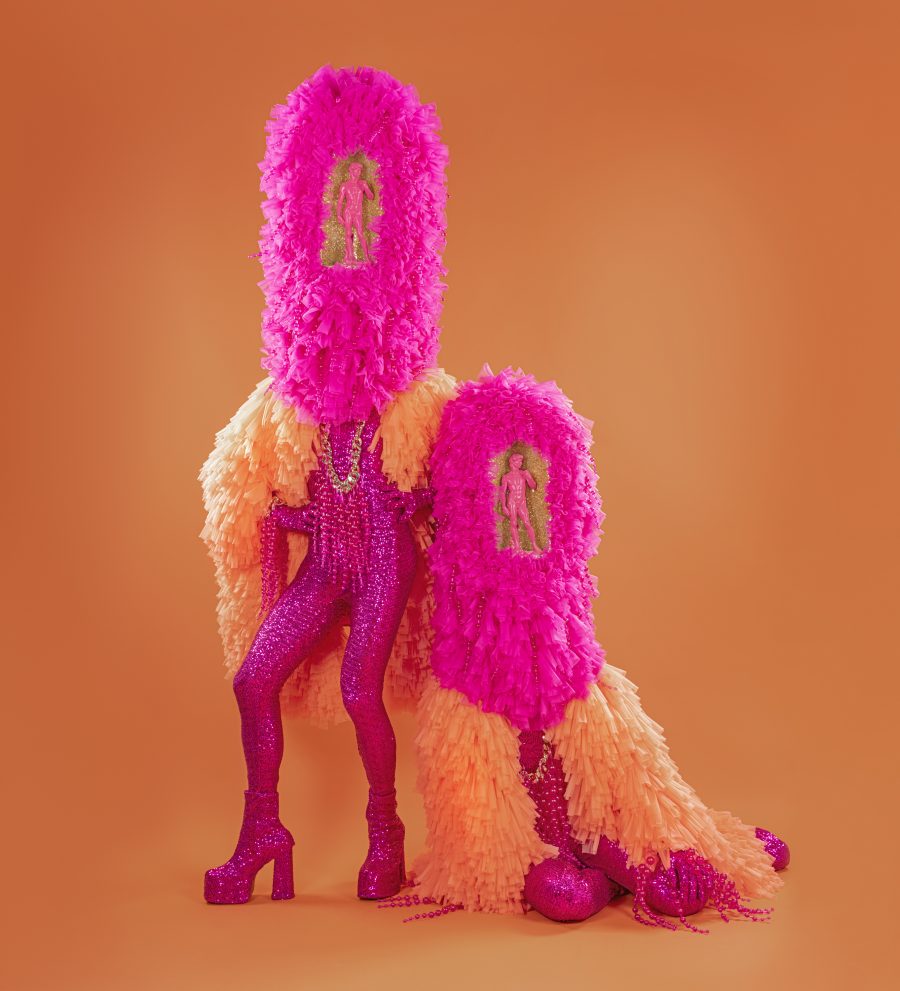 The Huxleys in frilly orange and pink outfits, with glittery pink bodysuits underneath. Their heads are obscured by frilly pink sculptures that contained a pink statue of David.