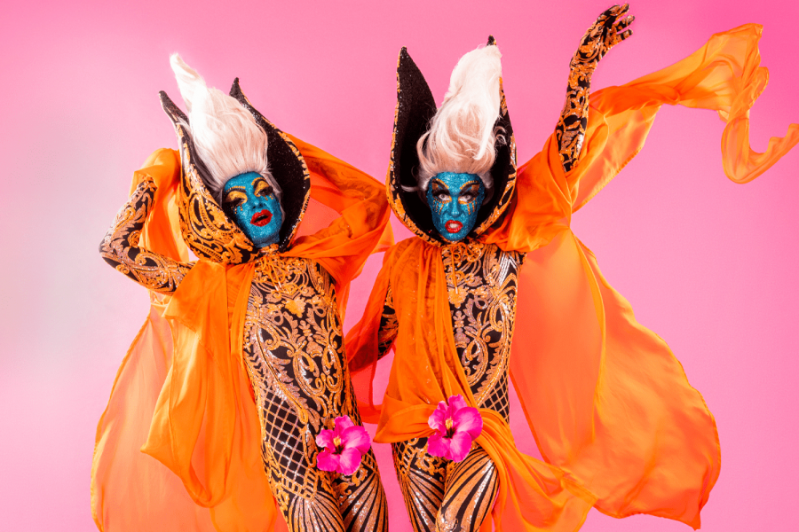 The Huxleys striking a pose with glittery blue faces and red lips, orange capes and beehive wigs. They have gold adorned jumpsuits and flowers on their crotches.