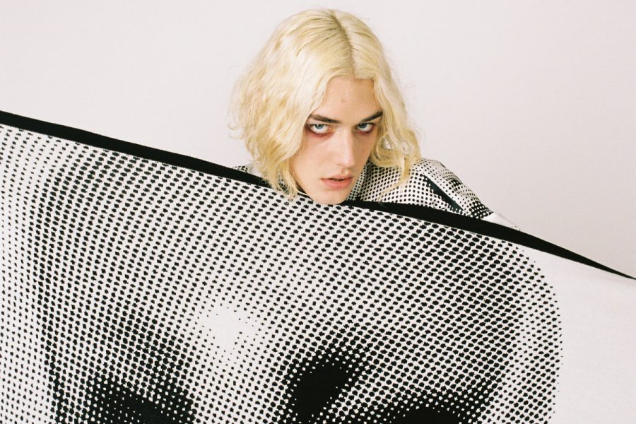 A person staring at the camera and wearing an outfit with a Ben Day-style dot pattern. They're holding fabric featuring a baby head printed in the same style. This design is by the label Jimmy D.