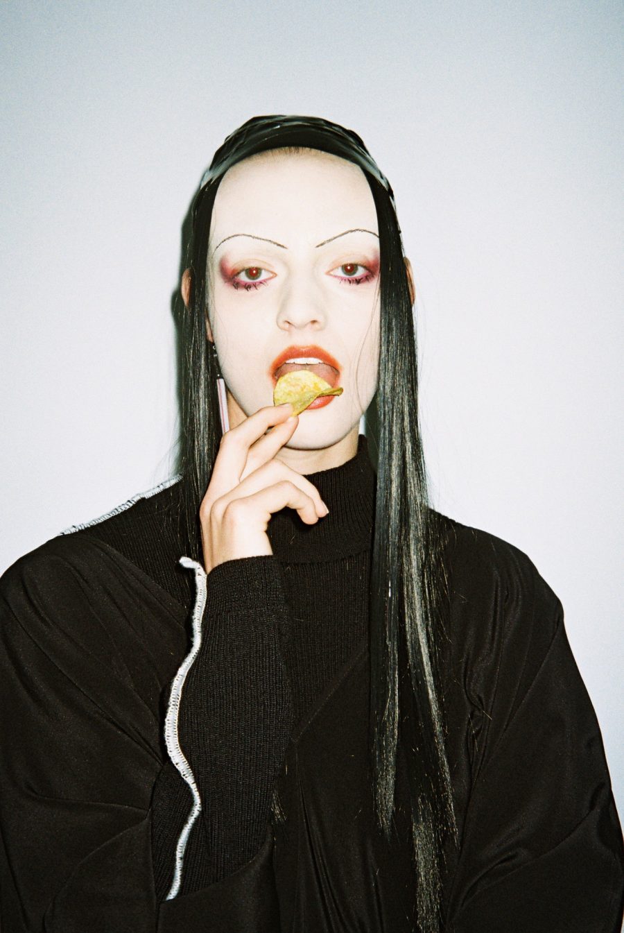 A person eating chips with a full face of makeup and a black sweater with white visible seams. Design by Jimmy D.