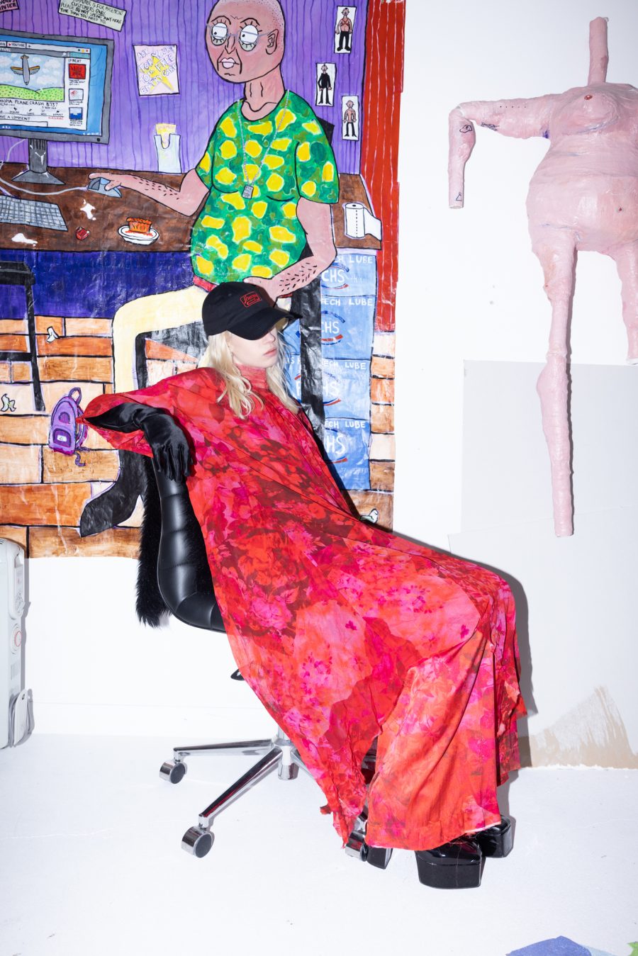 A person in a cap, red floral dress and platform shoes. They're sitting on a desk chair in front of some art.