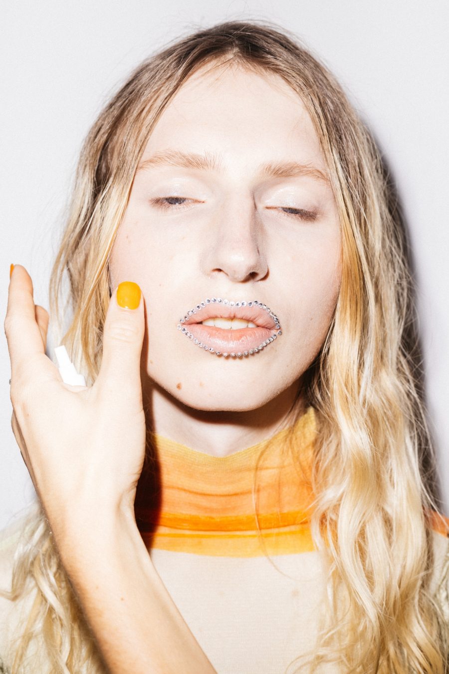 A close-up of a person wearing an orange top, orange nail polish and gems around their lips.