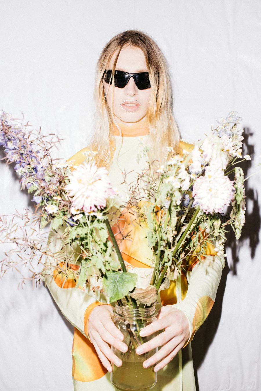 A person wearing sunglasses and an orange dress. They're holding flowers.