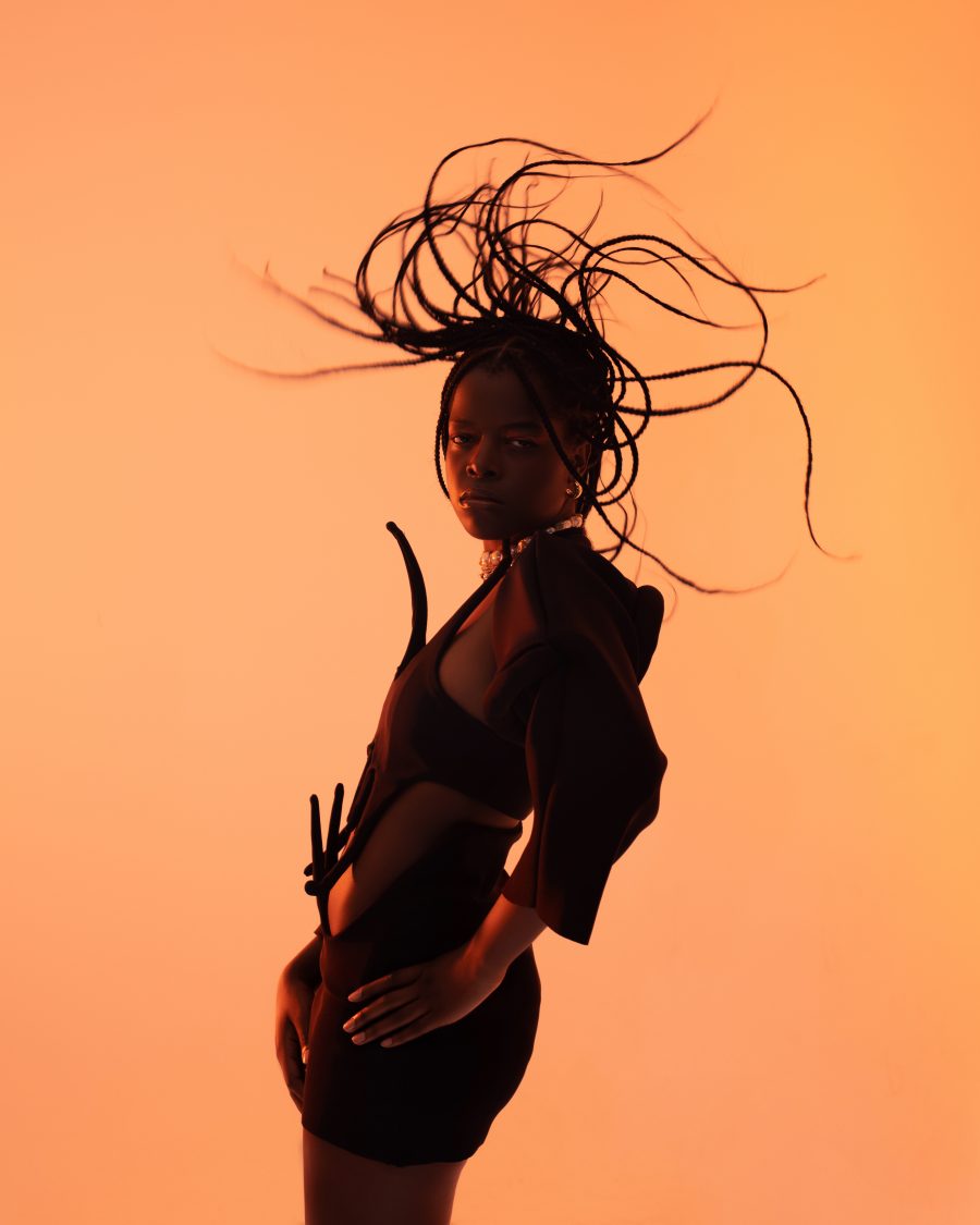 A person on an orange background with their braids flicking upwards.
