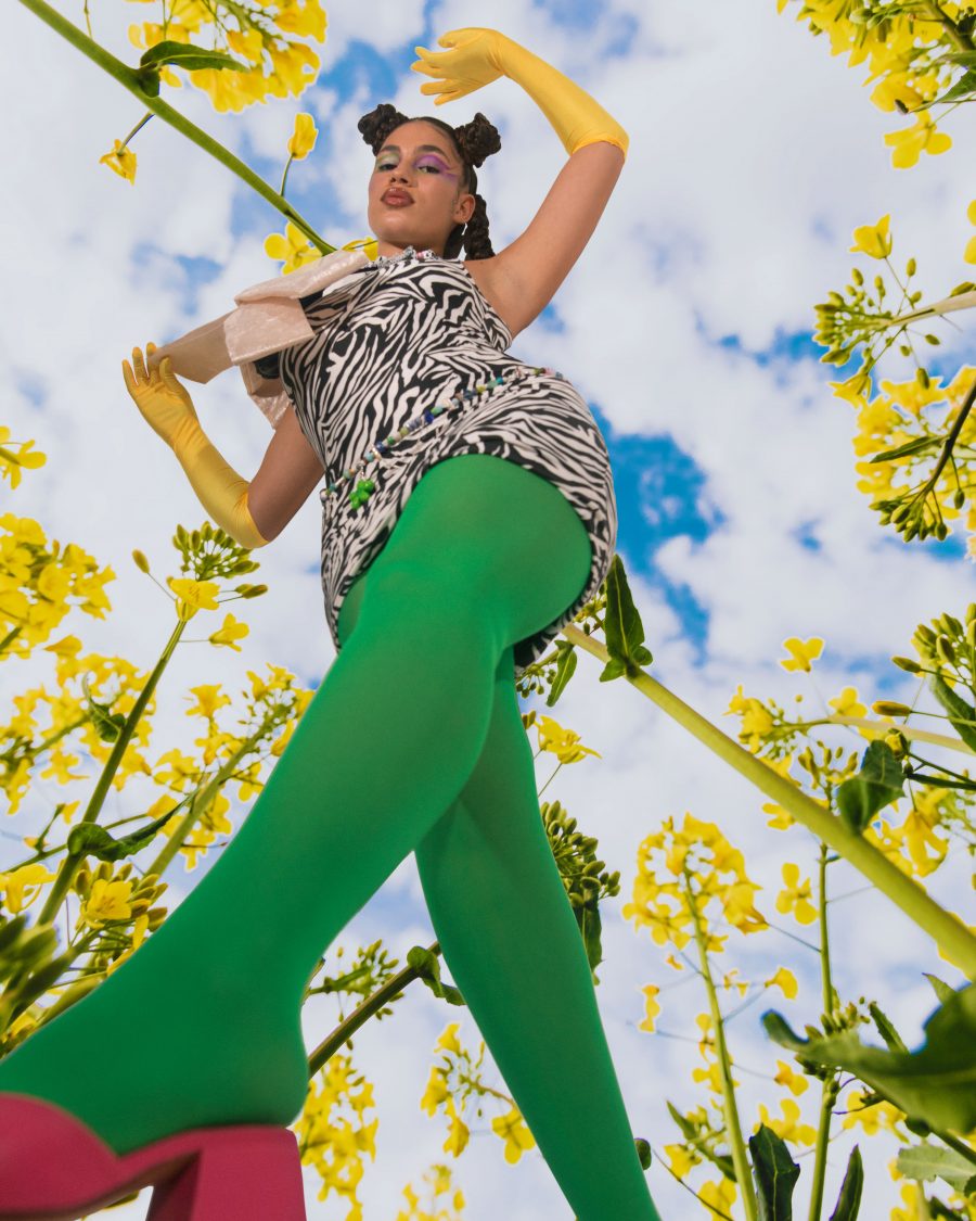 A person in green tights looking down at the camera, with sky and flowers behind them. Taken by Jade Florence.