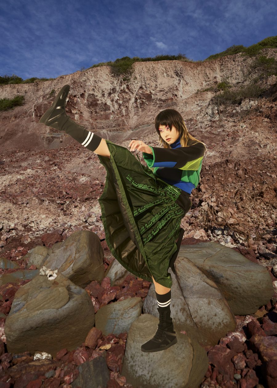 A person doing a high kick in a green and blue outfit, in front of a rocky background.