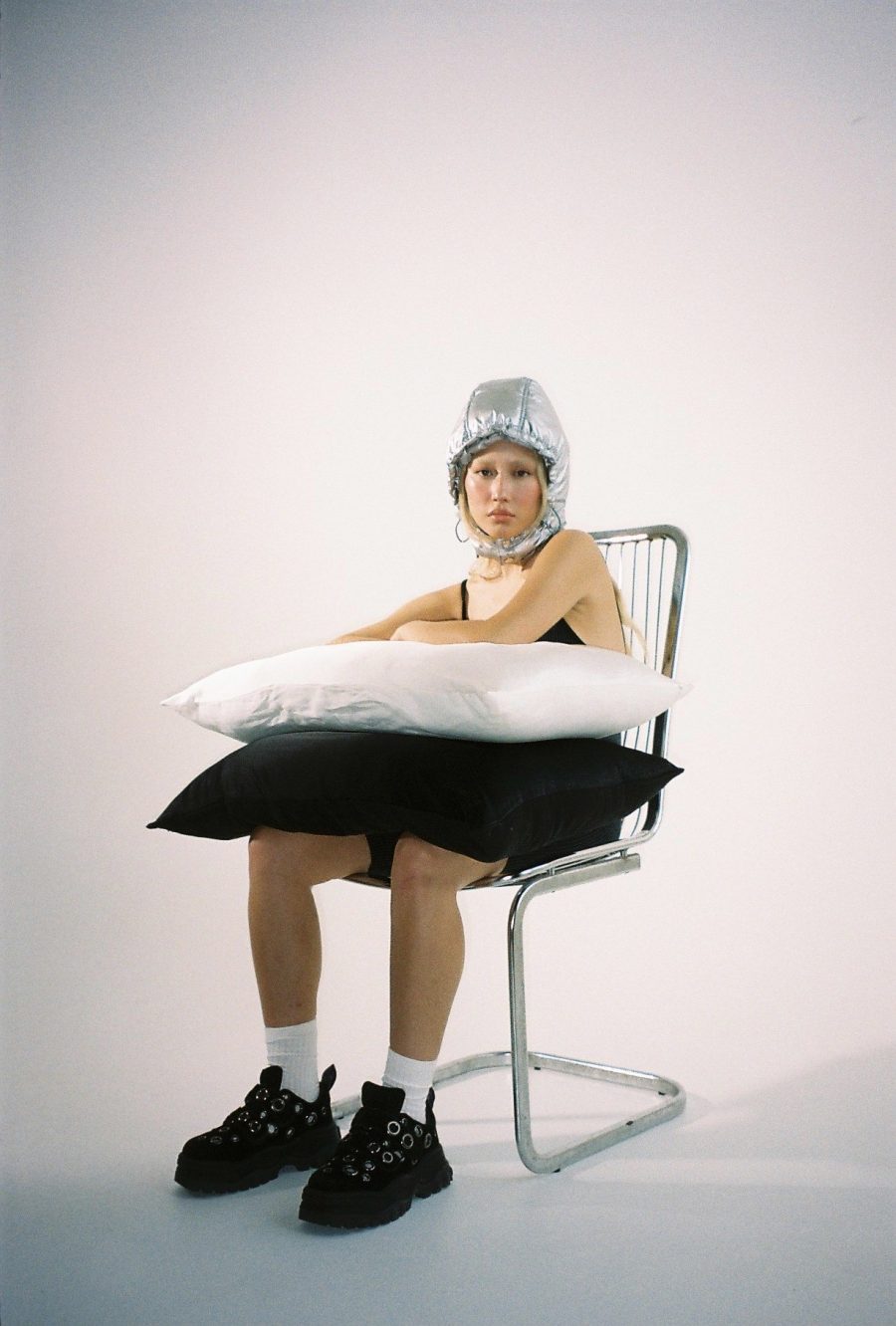 A person sitting on a metal chair holding pillows and wearing a silver hood. Taken by Lexi Laphor.