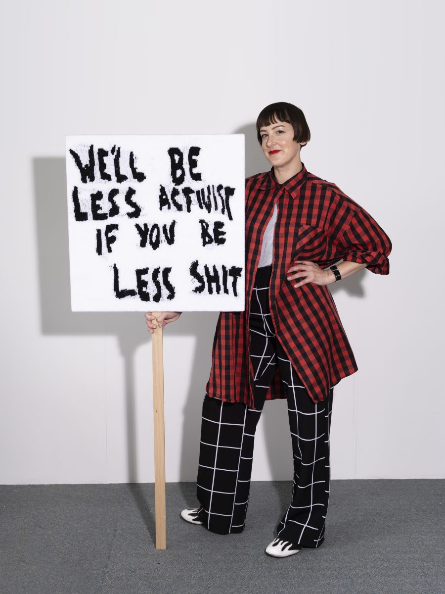 Kate Just holding her sign textile that says "WE'LL BE LESS ACTIVIST IF YOU BE LESS SHIT".