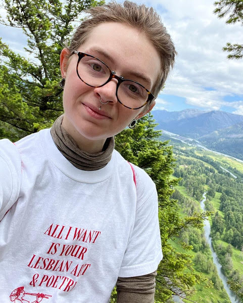 Headshot of Cavar, who wears glasses and a shirt that says "All I want is Your Lesbian Art & Poetry".