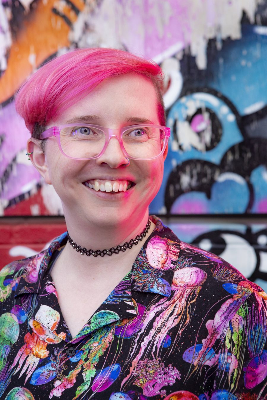 Rae White, a person with pink hair and glasses, smiling widely.