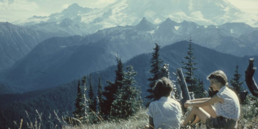 Two women sit chatting in a field, with mountains in the background.