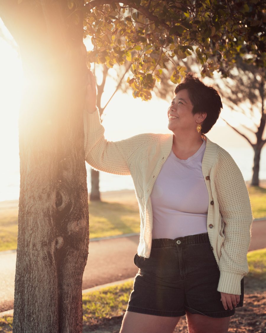 Kaya Ortiz standing next to a tree smiling, with sunlight peeking through behind them